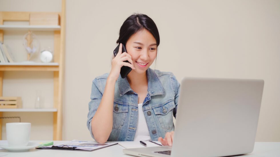 Business woman working on laptop and talking on VoIP phone while sitting on table in creative office.